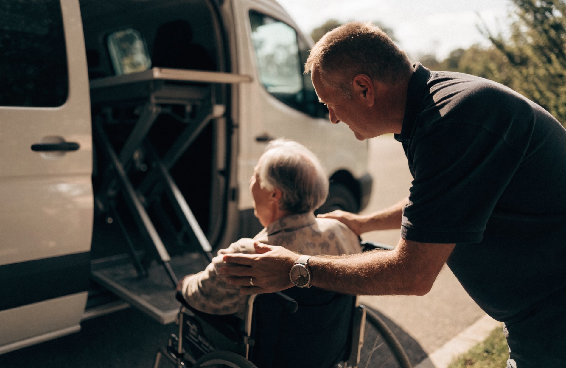 A non-emergency medical transport driver gently helping an older adult passenger board a clean, wheelchair-accessible van
